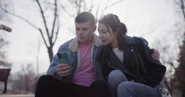 Genç bir çift parkta birlikte oturur, telefona bakar ve gülümserler. Mutlular ve birbirlerine eşlik etmekten zevk alıyorlar..
