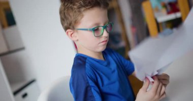 Gözlüklü genç bir çocuk masasına oturur ve yatak odasında kağıt uçak yapmak için bir kağıt parçasını dikkatlice katlar..