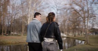 Genç bir çift, küçük bir nehrin kıyısındaki parkta el ele yürür, birlikte sakin bir gezintinin keyfini çıkarırlar..