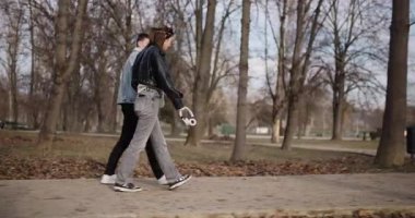 Genç bir çift, o bir çift kulaklık tutarken ağaçların arasındaki yolda el ele tutuşup parkta geziniyor..