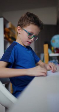 Gözlüklü genç bir çocuk masaya oturur ve bir parça kağıt katlar. Dikkatle bir origami figürü yaratırken odaklanmış ve kararlı..