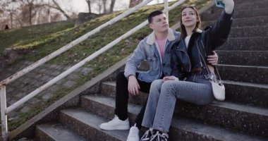 Genç bir çift parkta merdivenlerde selfie çeker. Gülümsüyorlar, poz veriyorlar ve başparmaklarını uzatıyorlar. Birlikte geçirdikleri zamanın tadını çıkarıyorlar..