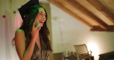 Mezuniyet şapkası ve cübbesi takmış genç bir kadın telefonda konuşuyor ve mezuniyet partisinde arkadaşlarıyla selfie çekiyor. Arkadaşları ona tezahürat yapıyor ve başarısını destekliyorlar. Arkadaşlığını gösteriyorlar..