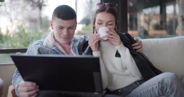 Genç bir çift bir kafede kahve içerken dizüstü bilgisayarlarını paylaşıyor ve birlikte gülüyorlar..
