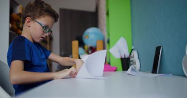 Gözlüklü genç bir çocuk odasında masasına oturur ve origami çalışırken bir parça kağıdı katlar..