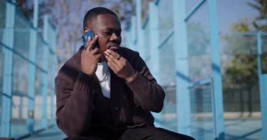 Genç Afrikalı Amerikalı bir adam bir telefon görüşmesi sırasında bankta oturur, spor salonunda bir şeyler anlatır ve el kol hareketi yapar..