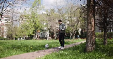 Bir adam şehir parkında tasmalı küçük köpeğiyle rahatça dolaşmaktan hoşlanır. Parlak güneş ışığı ağaçların arasından süzülür bahar çiçekleri ferahlatıcı bir atmosfer yaratır..