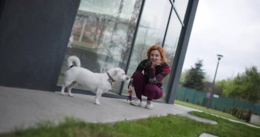 Kızıl saçlı bir kadın çömelmiş ve büyük pencereli modern bir binanın yakınında tasmalı beyaz bir köpekle etkileşime geçiyor. Açık hava ortamında birbirlerine eşlik etmekten zevk alıyor gibi görünüyorlar..