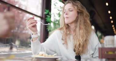 Uzun saçlı bir kız restoranda bir masada oturup yemeğinin tadını çıkarıyor. Güneş ışığı geniş pencerelerden akar ve sıcak ve davetkar bir atmosfer yaratır..