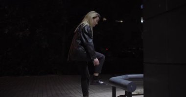 Loş bir açık hava ortamında, genç sarışın bir kadın çoraplarını bir park bankının yanında ayarlarken yakalanır..