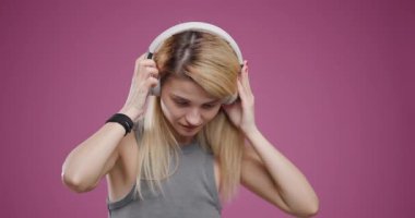 Gri kolsuz bluz ve beyaz kulaklıklar takan sarışın genç bir kadın, parlak pembe arka planda müziğin tadını çıkarıyor..