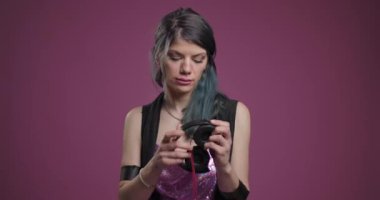 Mavi çizgili saçları olan düşünceli bir kadın pırıl pırıl giyinmiş, göz alıcı pembe bir arka planda kulaklıklarının kablosunu değiştiriyor..