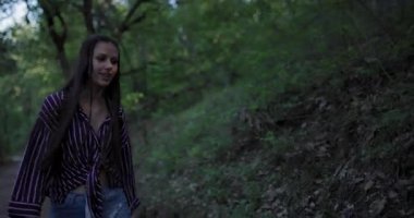 Genç bir kadın orman yolunda çizgili bluz ve şortla rahatça dolaşır. Kaygısız görünüyor, hoş bir günde çevreleyen yeşilliğin sükunetini kucaklıyor..