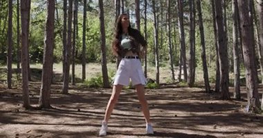 Genç bir kadın huzurlu bir orman ortamında enerjik bir şekilde dans ediyor. Uzun ağaçlar yaprakların arasından süzülen güneş ışığı sayesinde canlı bir atmosfer yaratıyor..