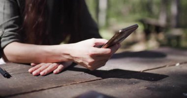 Bir insan sakin doğal ortamların ortasında, kırsal bir ahşap masada rahatlar, akıllı telefonunu kontrol eder ve açık havanın huzurlu atmosferinin tadını çıkarır..
