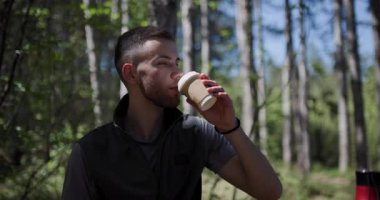 Genç bir adam elinde bir fincan kahveyle bir ormanda duruyor ve sakin atmosferi içine çekiyor. Güneş ışığı ağaçların arasından süzülüp, dışarıda huzurlu bir deneyim yaratıyor..