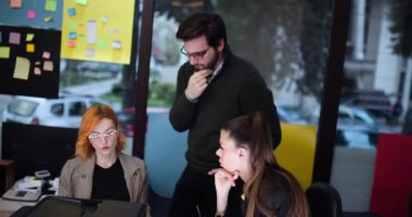 Üç kişi bir ofiste bilgisayar başında projeleri tartışmak için toplanıyor. Duvardaki notlara bakarken fikirleri paylaşıyor ve belgeleri gözden geçiriyorlar. Etkileşim canlı ve odaklıdır..