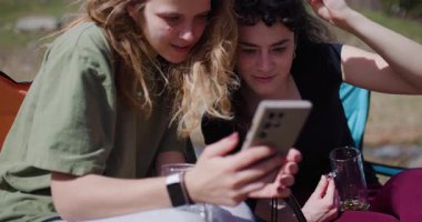 İki genç kadın derenin kenarındaki kamp sandalyelerinde rahatlar, içecekten zevk alır ve birlikte akıllı telefona bakarlar..