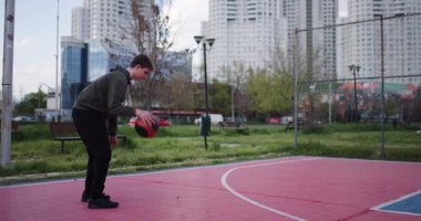 Genç bir çocuk dışarıda basketbol antrenmanı yapıyor, top sürüyor ve top atıyor. Egzersiz ve temiz havayı seviyor..