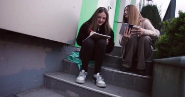 İki genç kız açık havada bir tablet ve defter kullanarak birlikte okuyorlar. İşbirliği yapıp öğreniyorlar. Arkadaşlık ve akademik odaklanma gösteriyorlar..