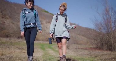 İki bayan arkadaş, sırt çantaları ve rahat giysiler giyerek doğa yürüyüşünden zevk alıyorlar. Birlikte yürür ve konuşurlar, temiz havanın ve manzaranın tadını çıkarırlar..