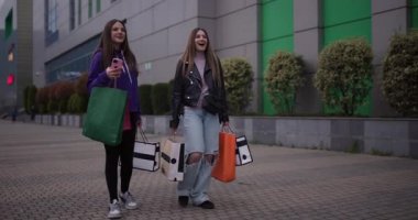İki mutlu genç kız alışveriş merkezinden ellerinde bir sürü alışveriş torbalarıyla çıkıyor, akıllı telefon kullanırken neşeyle konuşuyor ve gülümsüyorlar..