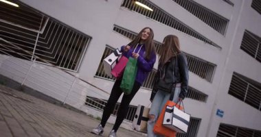 İki genç kız ellerinde alışveriş çantalarıyla dışarıda durup alışverişlerini tartışıyor ve birlikte vakit geçiriyorlar..