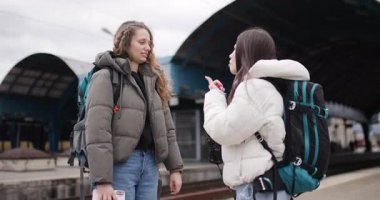 Sırt çantalı iki genç kadın trenin gelmesini beklerken bir tren istasyonunda konuşuyor ve gülüyorlar..