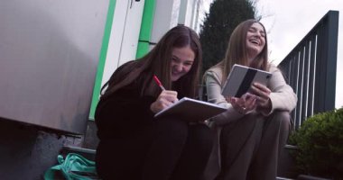 İki genç kız dışarıda ders çalışıyor. Bir kız deftere bir şeyler yazarken, diğeri tablette bir şeyler gösteriyor..