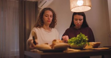 İki genç kadın mutfak masasında sandviç yapıyor. Yemeklerini hazırlarken konuşup gülüyorlar..