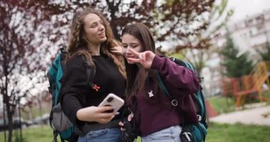 Sırt çantalı iki genç kadın parkta selfie ve fotoğraf çekiyor. Görüntüleri tartışıp birlikte gülüyorlar..