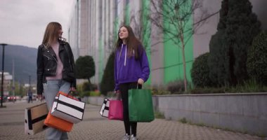 İki mutlu genç kız alışveriş yaptıktan sonra ellerinde renkli alışveriş torbalarıyla sokakta yürür ve gülümserler..