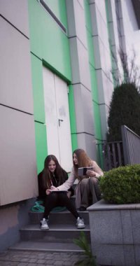 İki genç kız, bir defter ve tablet kullanarak modern bir binanın dışında birlikte okuyorlar. İşbirliği yapıp öğreniyorlar. Eğitim ve dostluğu somutlaştırıyorlar..