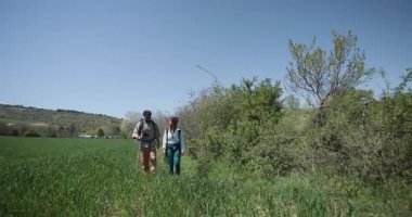 Güneşli bir günde bir erkek ve bir kadın tarlada yürüyüş yaparlar. Sırt çantası takıyorlar ve yürüyüş sopası kullanıyorlar..