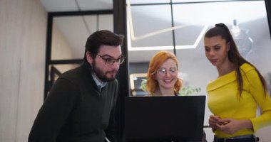 Modern bir ofiste dizüstü bilgisayarın etrafında üç kişi toplanıyor. Ekranda bir şey izlerken şaşkınlık ve ilgi gösteriyorlar. Ayar parlak ve doğal ışıkla dolu..