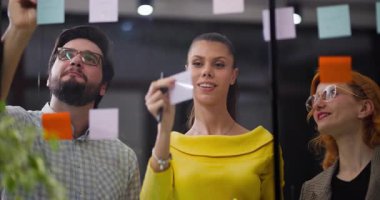 Grup, fikirleri paylaşmak ve projeleri tartışmak için ofiste toplanıyor. Düşünceleri özetlemek ve işbirliği yapmak için cam bir tahtaya yapışkan notlar koyarlar. Bu onların planlama sürecine yardımcı olur..