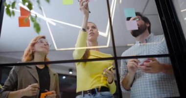 Bir grup insan çalışma masasında fikirlerini tartışıyor ve paylaşıyor, aynı zamanda ofiste cam bir duvara yapışmış renkli yapışkan notlarla etkileşimde bulunuyor. Birlikte yaratıcı bir sürece giriştiler..