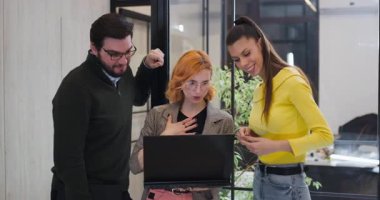 Üç kişi bir dizüstü bilgisayarın etrafında toplanıp fikirlerini paylaşıyor ve ofis ortamında bir projeyi tartışıyorlar. Çalışma saatleri boyunca parlak bir alanda iş birliği yaparken meşgul görünüyorlar..