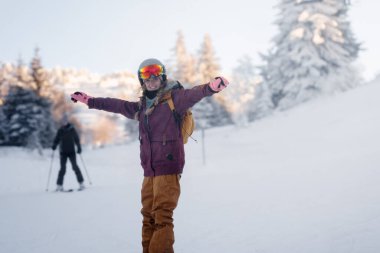 Bir insan karla kaplı bir dağda kayak yapmaktan zevk alıyor. Kollarını açıp gülümsüyorlar. Arka planda başka bir kayakçı var. Gökyüzü açık, ağaçlar karlı..