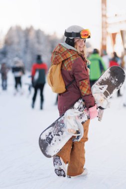 Sarı saçlı ve kasklı bir insan karlı bir alanda kar kaykayı taşır. Arka planda kış sporlarını ve parlak güneşin altındaki aktiviteleri seven bir grup insan var..
