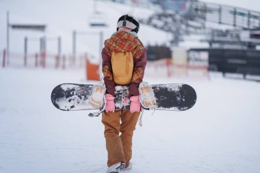 Kar kayakçısının elinde kayak merkeziyle karlı bir yolda tek başına yürüyor. Gün ışığında arka planda karla kaplı yamaçlar ve ekipmanlar görülebilir..
