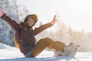 Bir snowboardcu kolları havada otururken karda vakit geçirmekten zevk alır. Güneş gökyüzünde ışıl ışıl parlıyor, etrafındaki karlı manzarayı ve ağaçları aydınlatıyor..