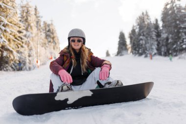 Karlı bir yerde kar kayağı yaparken kask ve eldiven takan bir kız. Ağaçlar ve diğer kış sporu meraklılarıyla çevrili kış gününün tadını çıkarıyor..