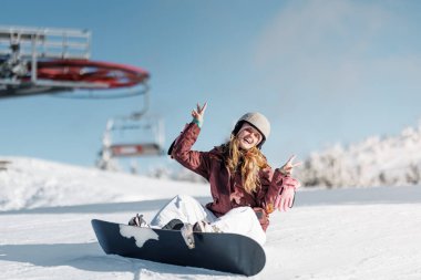 Bir kız kayak merkezindeki snowboard 'unda oturuyor. Barış işareti olarak elleri ile neşeli bir ifade sergiliyor. Gökyüzü parlak ve berrak, her tarafı karla çevrili ve arkasında bir kayak asansörü var..