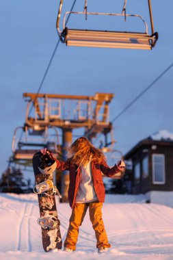 Genç bir insan karda elinde bir snowboard ile duruyor. Kayak asansörü arka planda turuncu bir gökyüzü ile birlikte. Kayak merkezindeki kış akşamına benziyor..