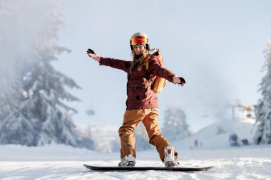 Kar yamacında kask ve eldiven takan bir snowboardcu. Arka planda, karlı ağaçlar ve kayak asansörleri var. Güneş parlak parlıyor. Açık bir kış günü..