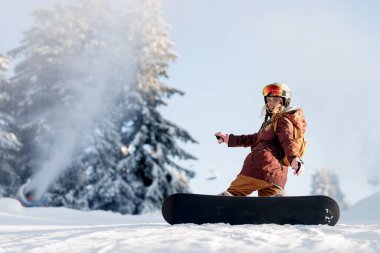 Karlı bir yamaçtan snowboard 'a binen biri. Arka planda karla kaplı ağaçlar görülüyor. Kayakçı kask ve koruyucu giysiler giyerken güneş parlıyor..