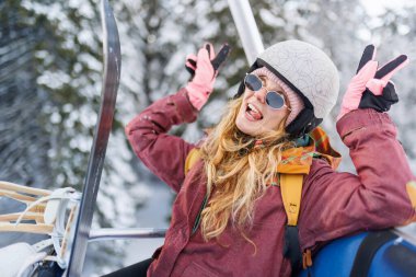 Bir kız dağlarda kayak asansörüne binerken eğleniyor. O, bir kask ve eldiven giyerken gülümsüyor ve elleriyle barış işaretleri yapıyor. Etrafındaki ağaçlar karla kaplı..