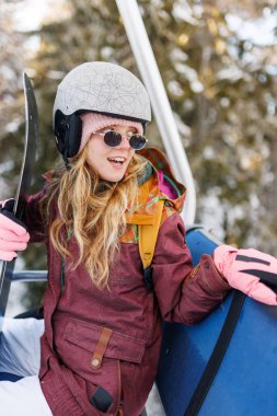 Kask ve güneş gözlüğü takmış, elinde kayak tahtası tutan bir kız. Karla kaplı ağaçlar arka planda kış manzarası yaratıyor. Yolculuğun tadını çıkarırken mutlu görünüyor..
