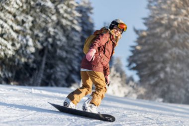 Kış ortamında bir kişi yamaçtan snowboard yapıyor. Parlak güneş ışığı karların ve etraftaki ağaçların üzerinde parlıyor. Snowboard 'cu kalın kıyafetler ve kask giyer..